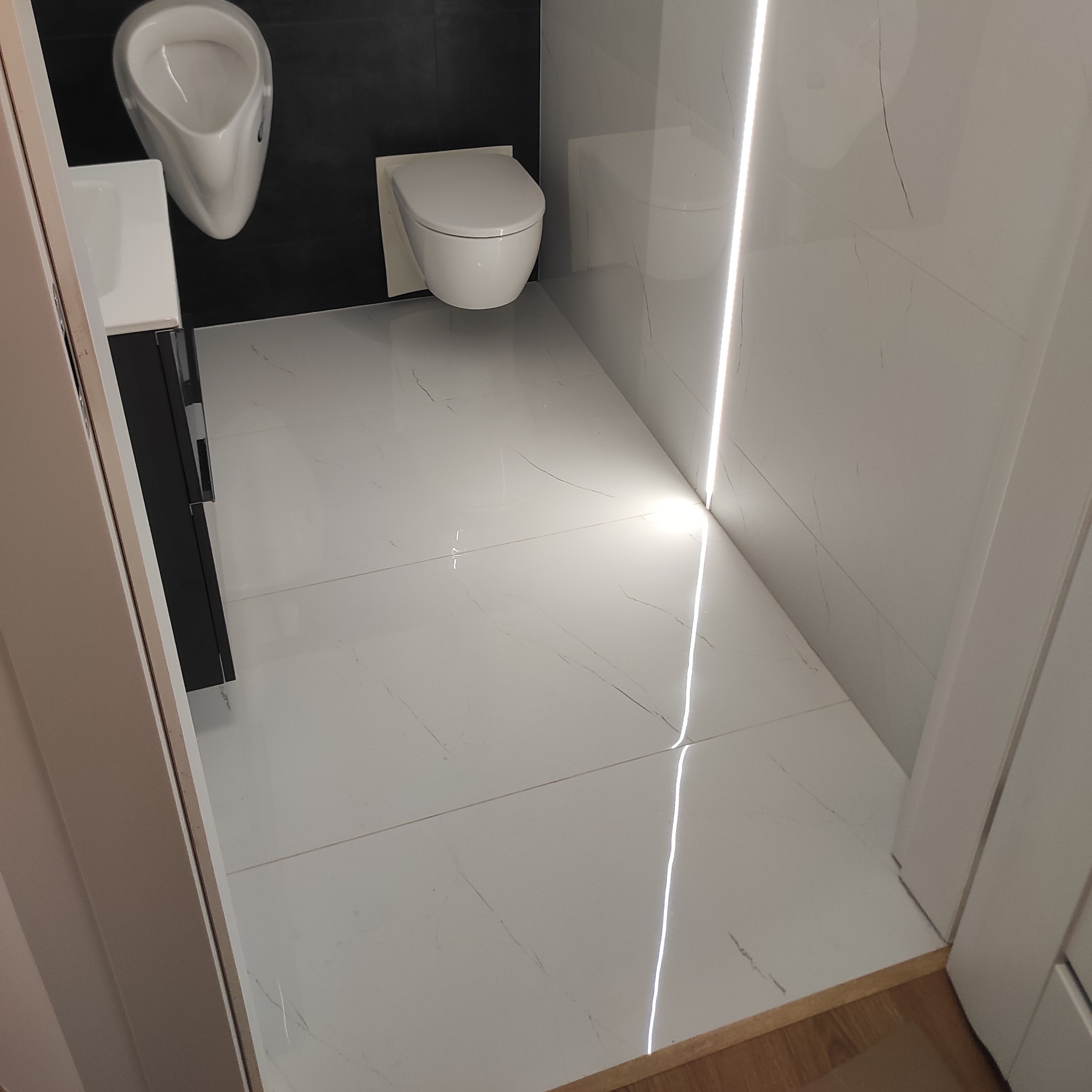 Nowoczesna toaleta z podwieszaną miską WC i pisuar, białe płytki imitujące marmur z podświetleniem LED w fugach, czarna ściana za pisuar.