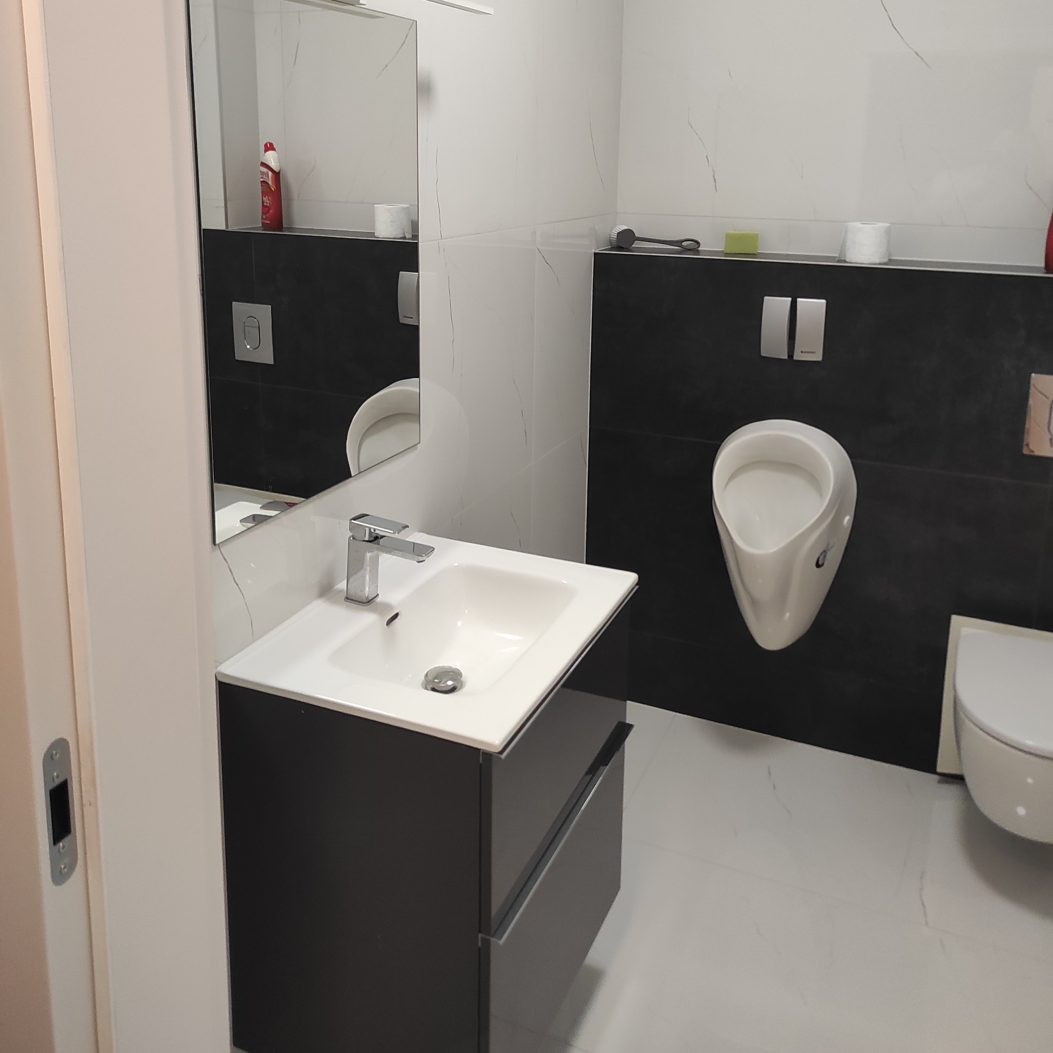 Nowoczesna łazienka z czarno-białymi płytkami imitującymi marmur, wiszącą umywalką z szafką w kolorze grafitowym, wiszącym pisuarzem i toaletą, odbicie butelki środka czyszczącego w lustrze.