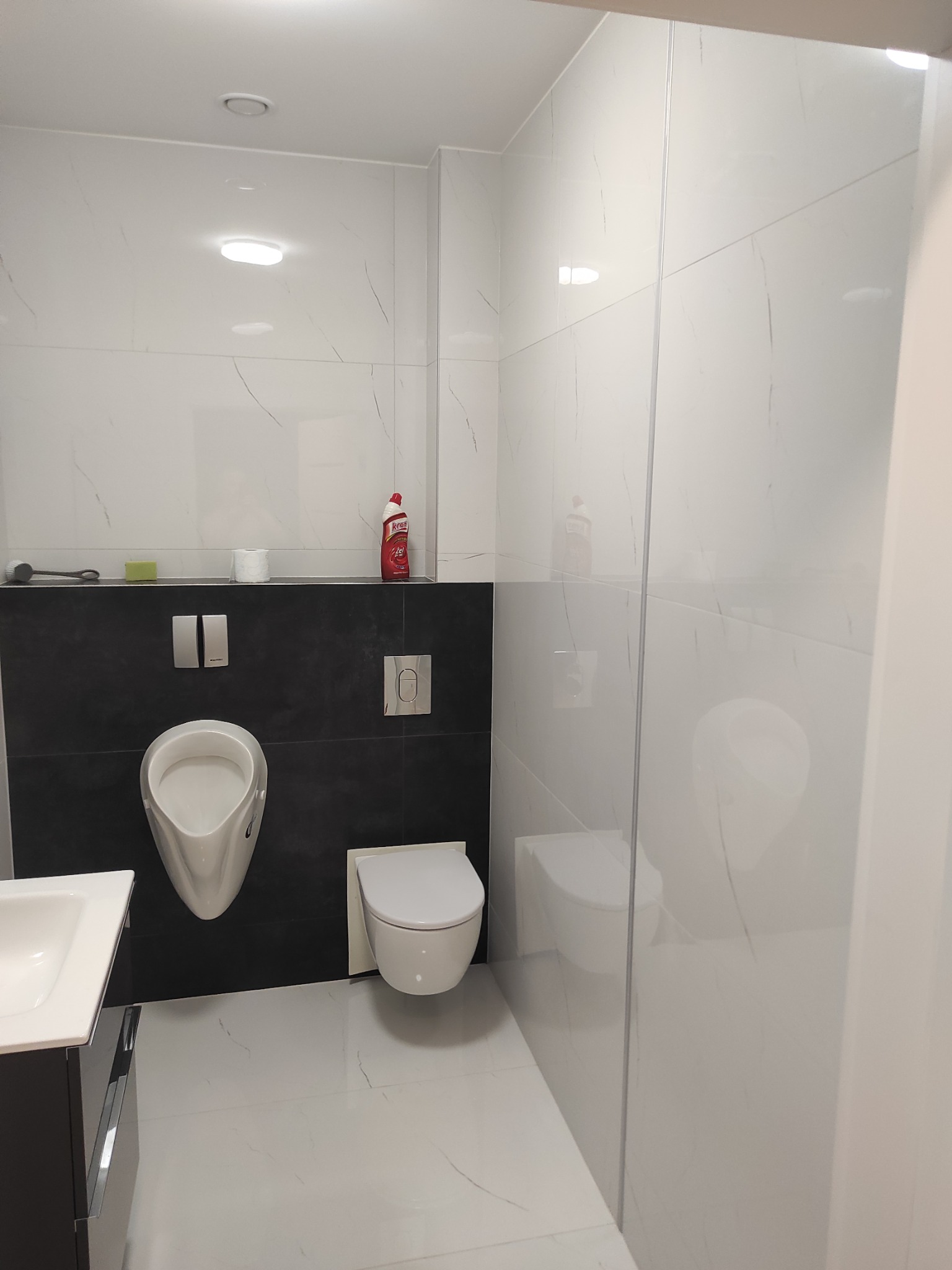 Nowoczesna łazienka z białymi marmurkowymi płytkami, czarną ścianą akcentującą, wiszącą miską WC, pisuar i umywalką z czarną szafką, widok przez szklany panel prysznicowy.