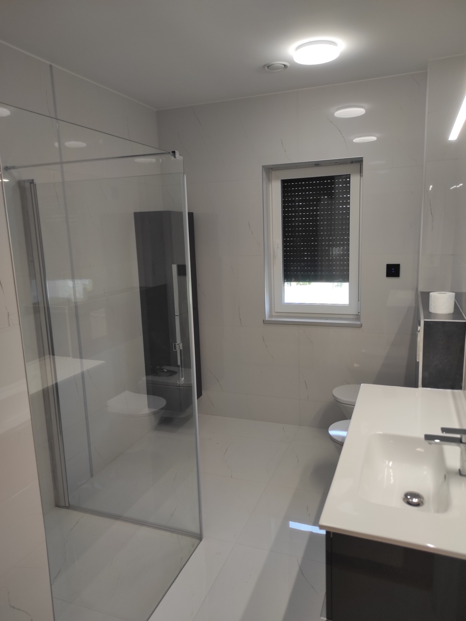 Nowoczesna, minimalistyczna łazienka z przeszkloną kabiną prysznicową, umywalką z szafką w kolorze grafitowym, toaletą podwieszaną i oknem z żaluzjami w białej ramie, wykończona płytkami...