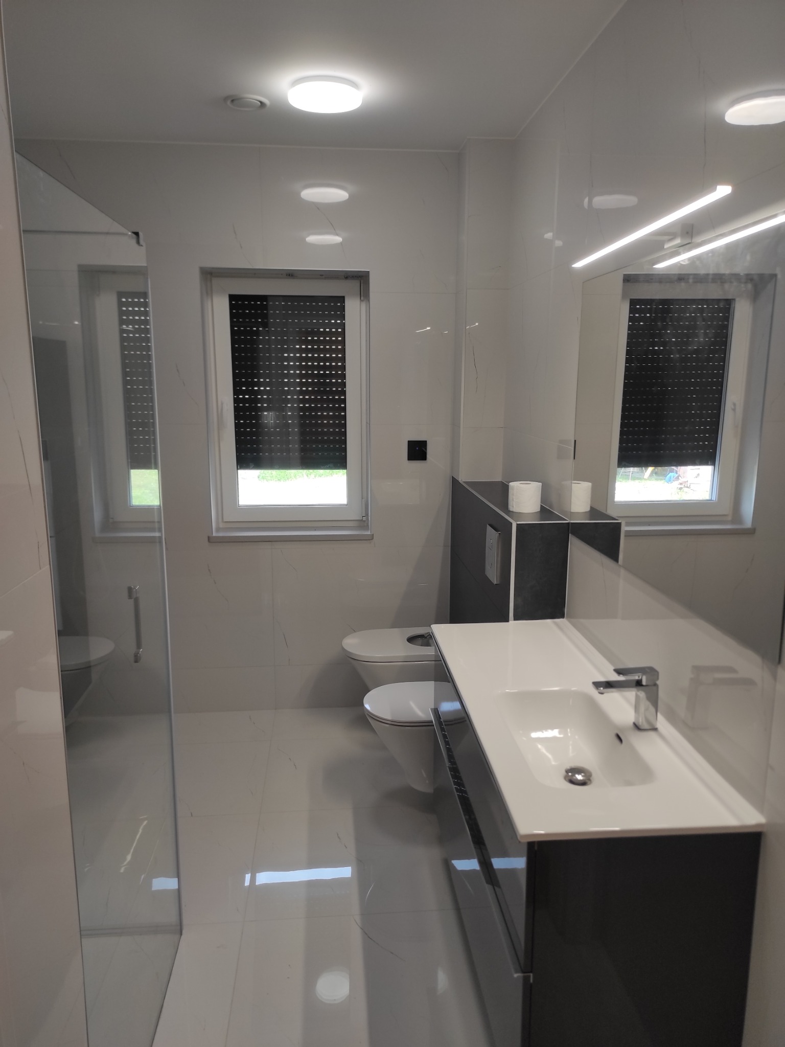 Nowoczesna łazienka w bieli i szarości z kabiną prysznicową typu walk-in, umywalką nablatową z szafką i toaletą podwieszaną z zabudową.