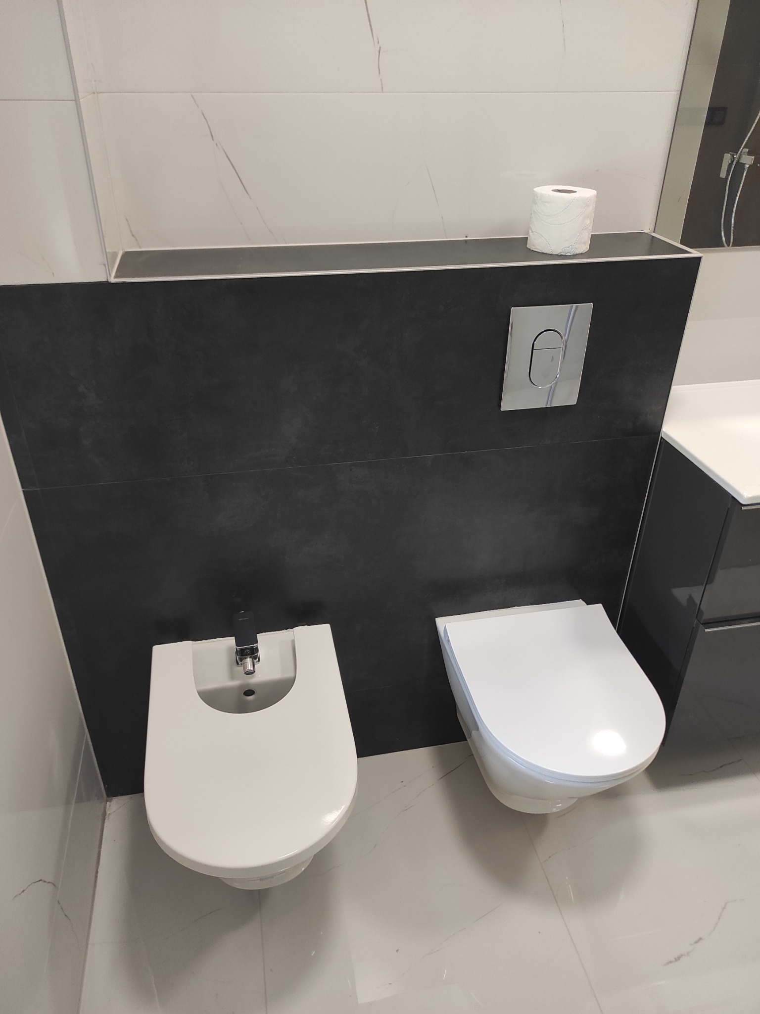 Nowoczesna łazienka z biało-czarnymi płytkami marmurkowymi, wiszącą toaletą i bidetem z chromowaną armaturą oraz przyciskiem spłukującym w kolorze srebrnym.