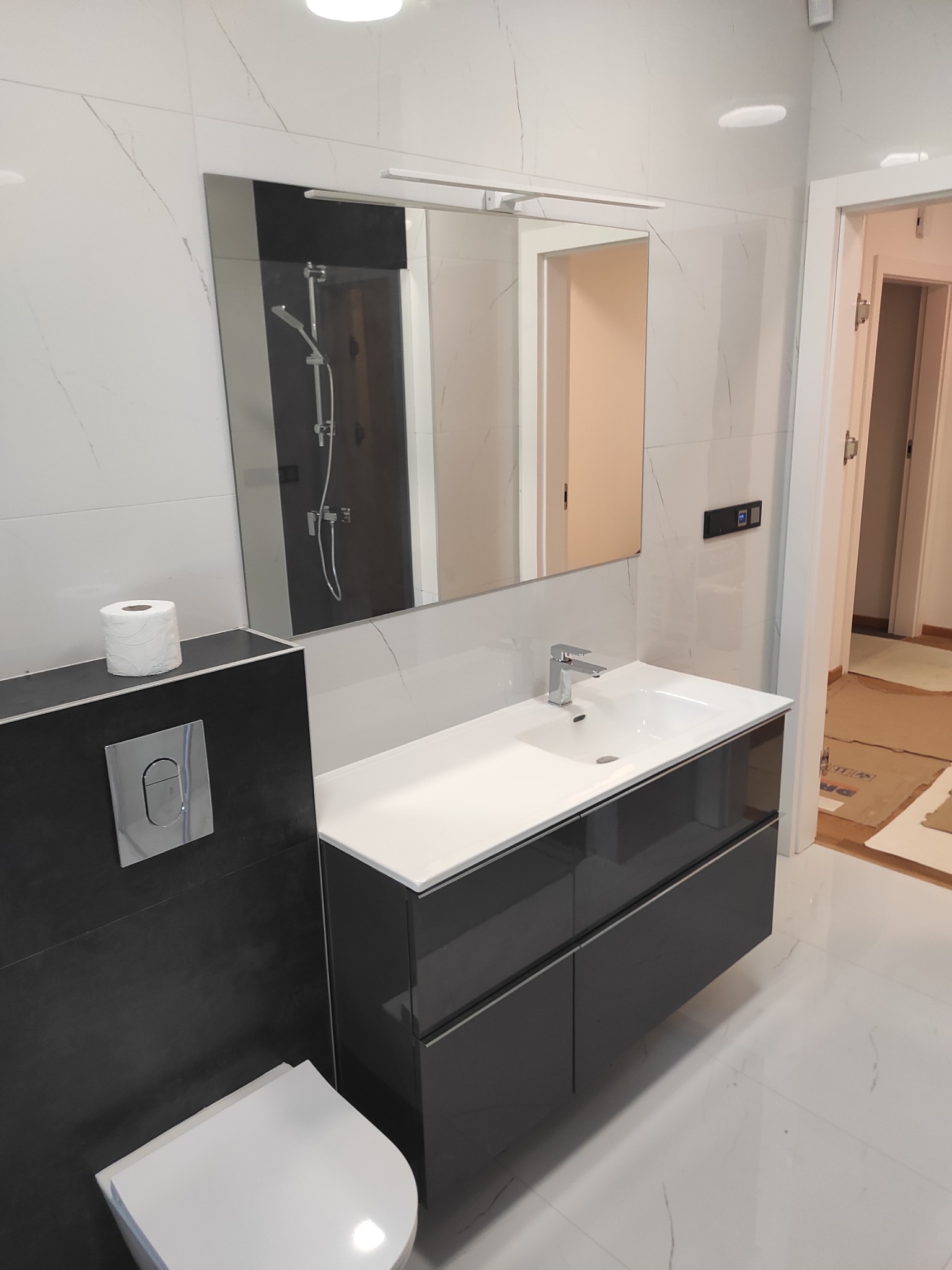 Nowoczesna łazienka z białymi marmurkowymi płytkami, szarą szafką pod umywalką, lustrem z oświetleniem LED i wiszącą muszlą klozetową z czarnym panelem. W tle widoczne drzwi do innego pomieszczenia...