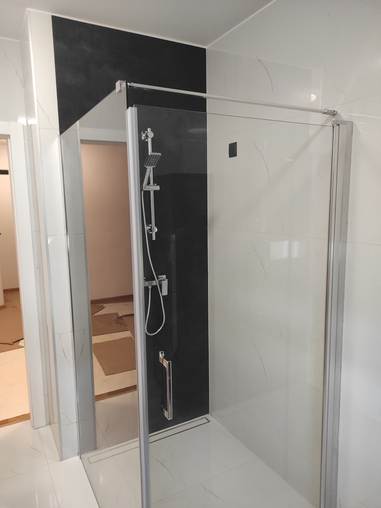Nowoczesna kabina prysznicowa z czarnym panelem, szklaną ścianą i chromowaną armaturą, widok z zewnątrz na białe płytki imitujące marmur i podłogę zabezpieczoną kartonem.