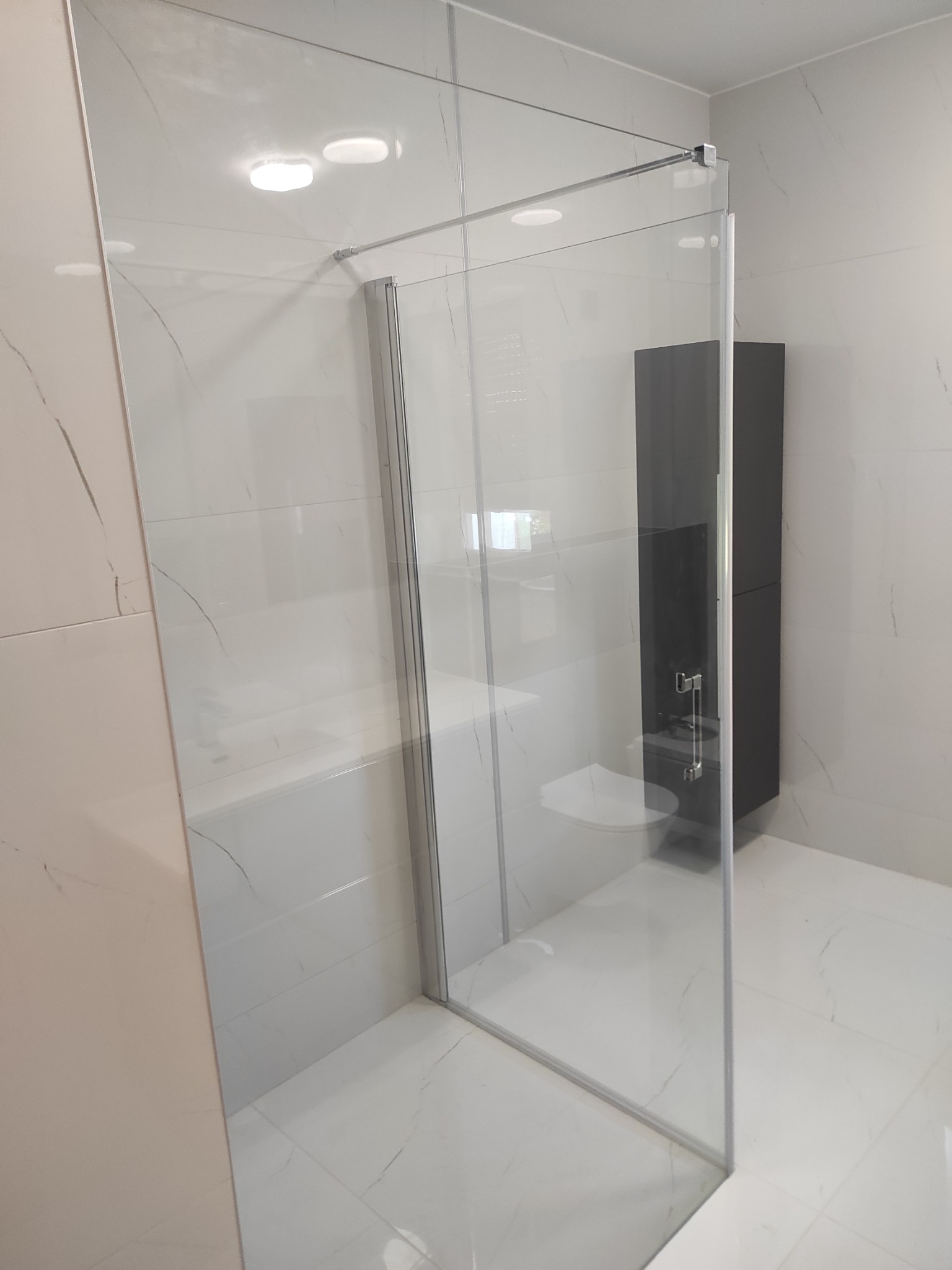 Nowoczesna kabina prysznicowa ze szklanymi ścianami, chromowanymi okuciami i minimalistycznym czarnym regałem w tle, na białych płytkach imitujących marmur.