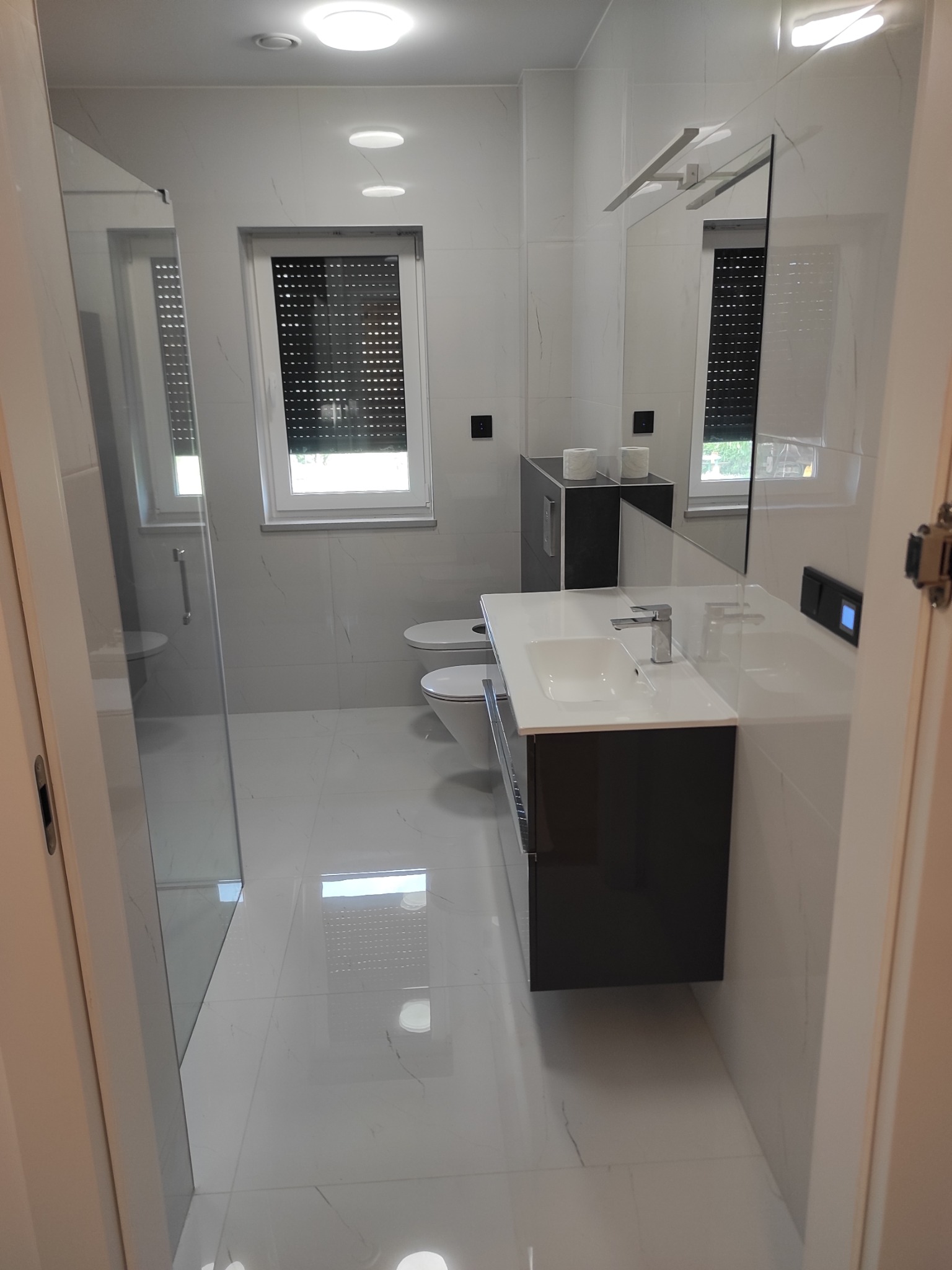 Nowoczesna, jasna łazienka z białymi marmurkowymi płytkami, czarnym blatem pod umywalką i kabiną prysznicową ze szkła hartowanego. W tle widoczne okno z żaluzjami i toaleta.