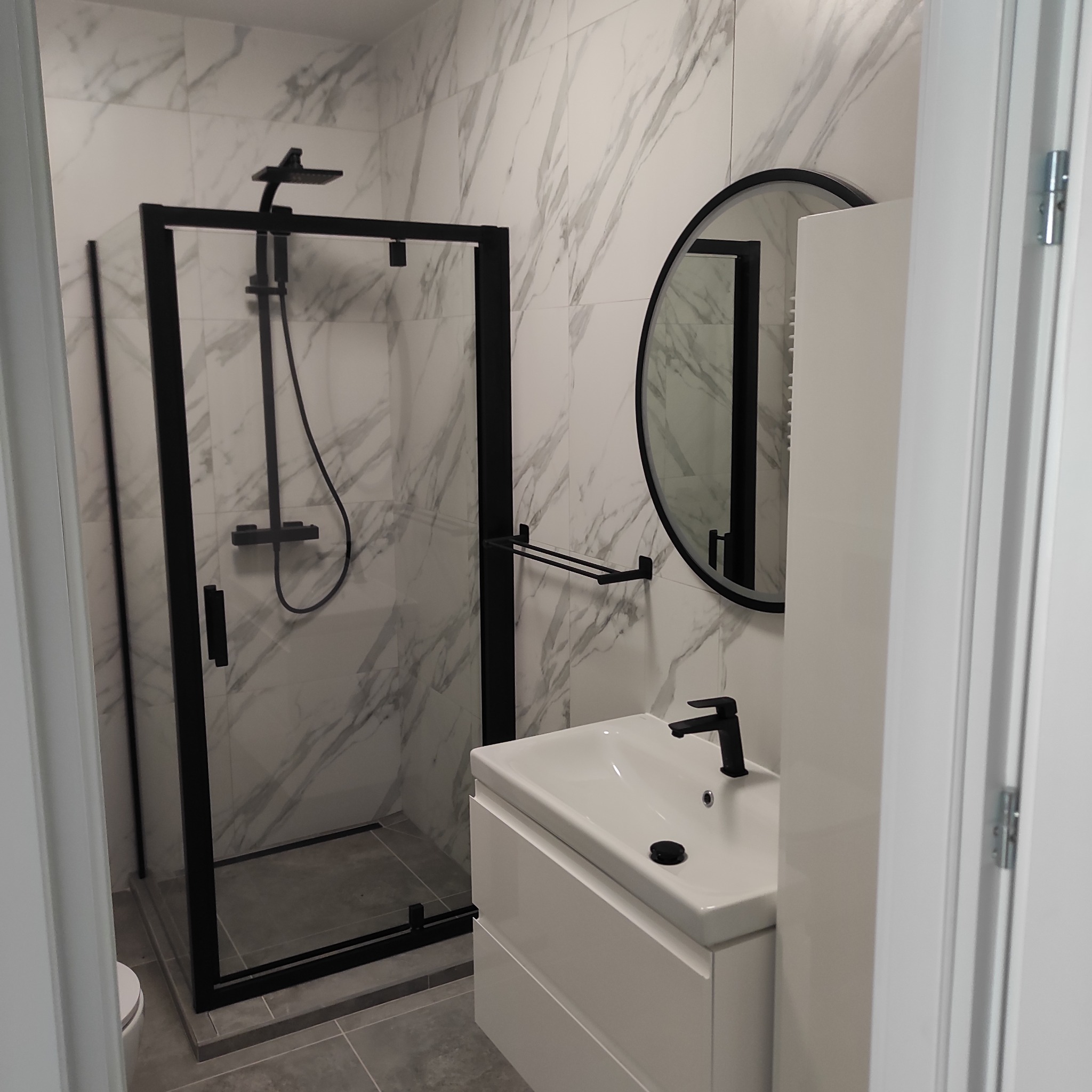 Nowoczesna łazienka z kabiną prysznicową typu walk-in z czarnymi profilami, okrągłym lustrem z czarną ramą i białą szafką podumywalkową z czarną armaturą, wykończona płytkami imitującymi marmur.