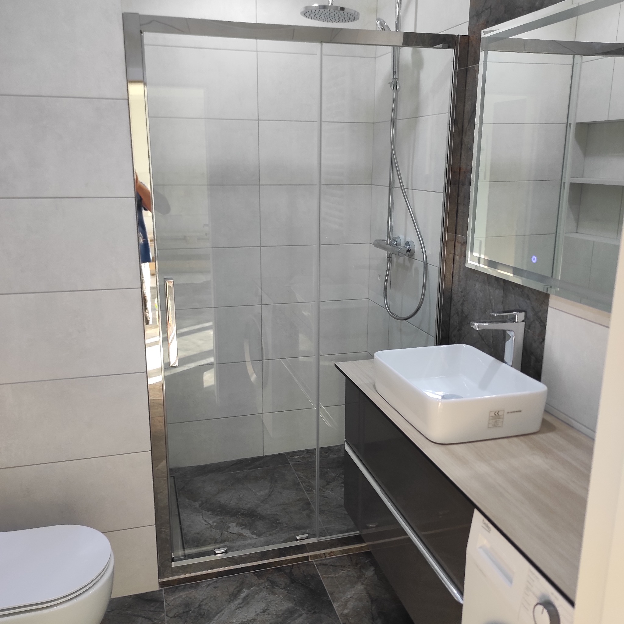 Nowoczesna łazienka z kabiną prysznicową typu walk-in, umywalką nablatową na szafce z szufladami i podświetlanym lustrem, wykończona szarymi płytkami imitującymi beton.