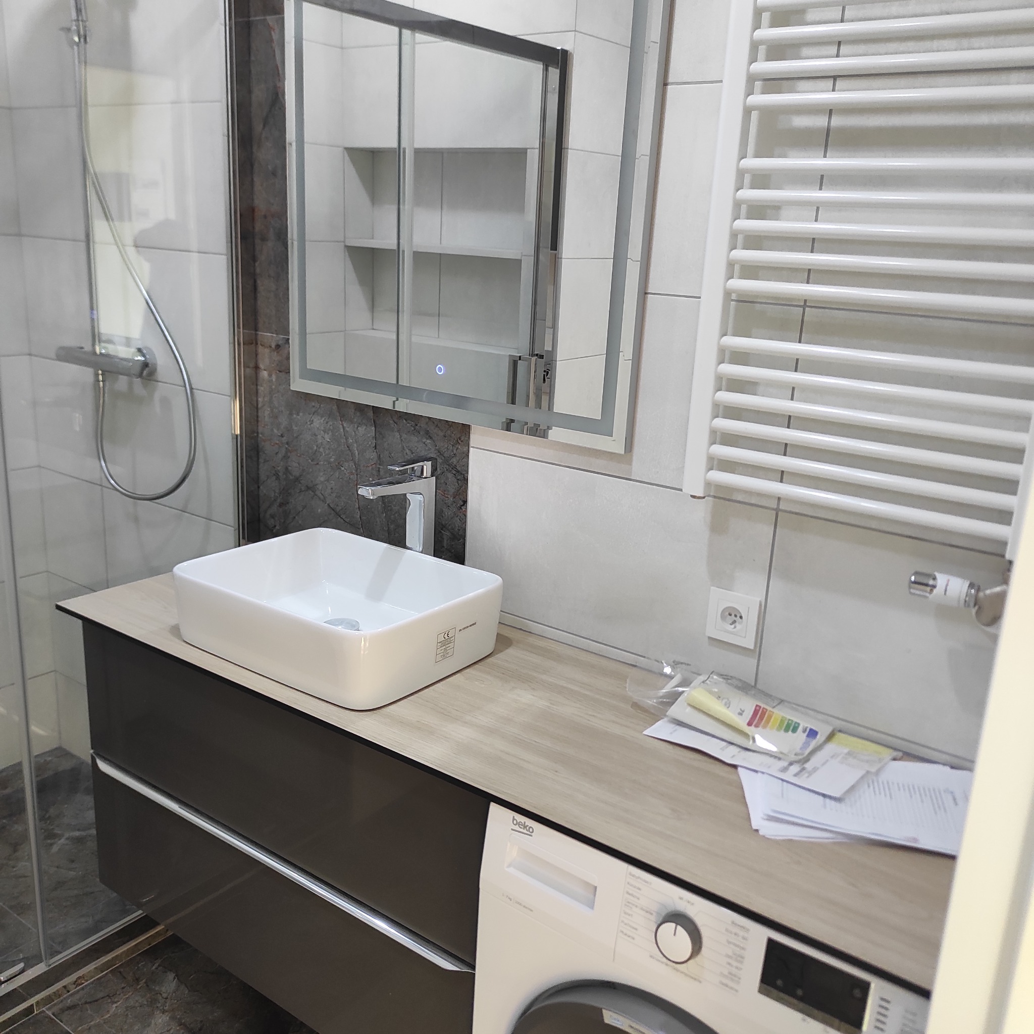 Nowoczesna łazienka po remoncie: biała umywalka nablatowa, bateria, podświetlane lustro z szafką, grzejnik drabinkowy, kabina prysznicowa i pralka Beko z dokumentacją techniczną.