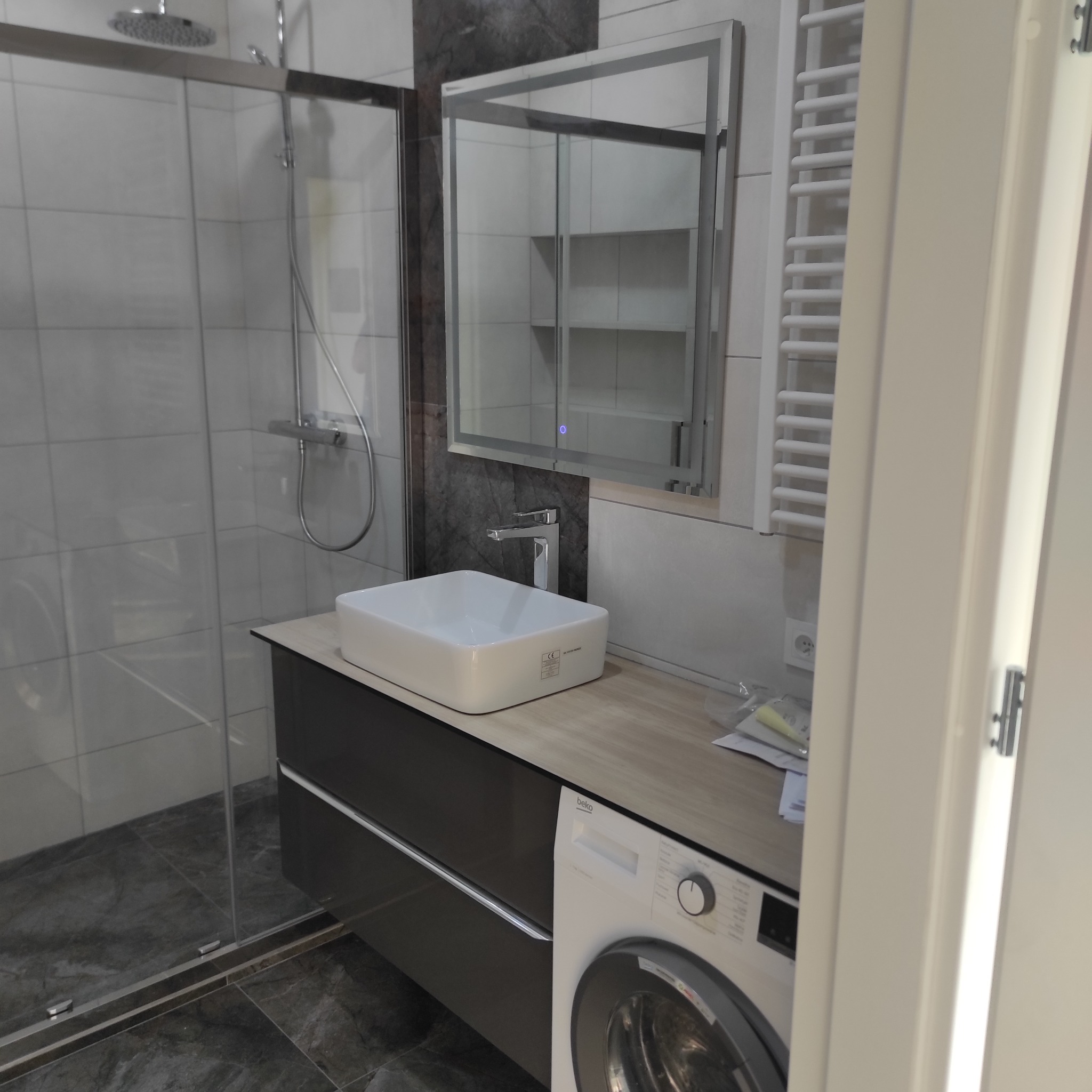 Nowoczesna łazienka z kabiną prysznicową, umywalką nablatową na szafce z szufladami i podświetlanym lustrem. Obok grzejnik drabinkowy i pralka. Widoczne marmurowe płytki na podłodze i ścianie.