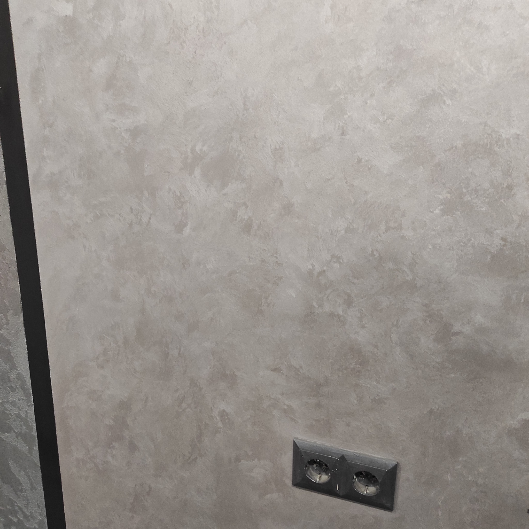 Ściana z dekoracyjnym, beżowym tynkiem strukturalnym i gniazdkiem elektrycznym w czarnej obudowie.