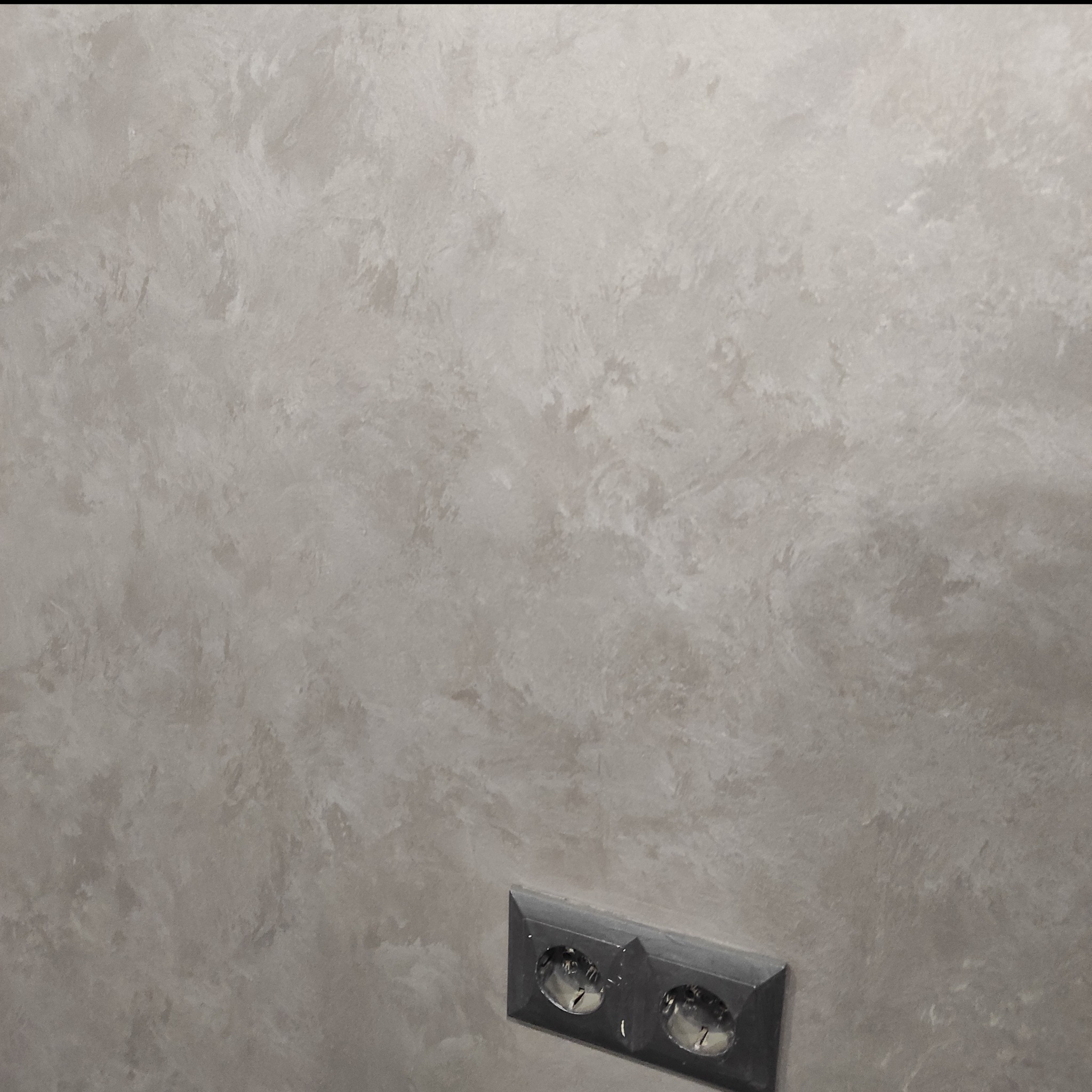 Ściana pokryta dekoracyjnym tynkiem w odcieniach szarości, z widocznym gniazdkiem elektrycznym w dolnym rogu.