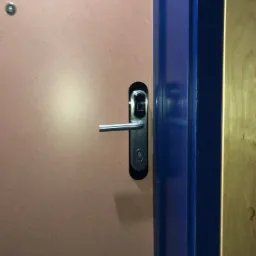 Zewnętrzny zamek elektroniczny z czytnikiem kart na brązowych drzwiach, widoczna niebieska ościeżnica i fragment drewnianej ściany w tle.