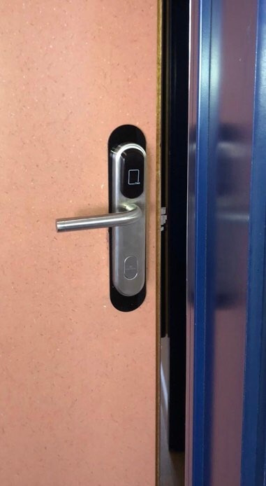 Zamek elektroniczny z czytnikiem kart na brązowych drzwiach wejściowych, widoczna klamka i fragment niebieskiej ościeżnicy.