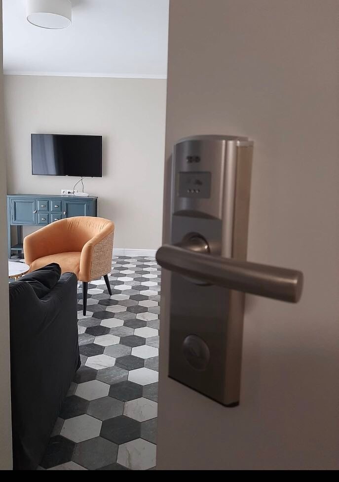 Elektroniczny zamek do drzwi wejściowych w apartamencie, widok z korytarza na salon z wzorzystą podłogą i meblami w stylu retro.