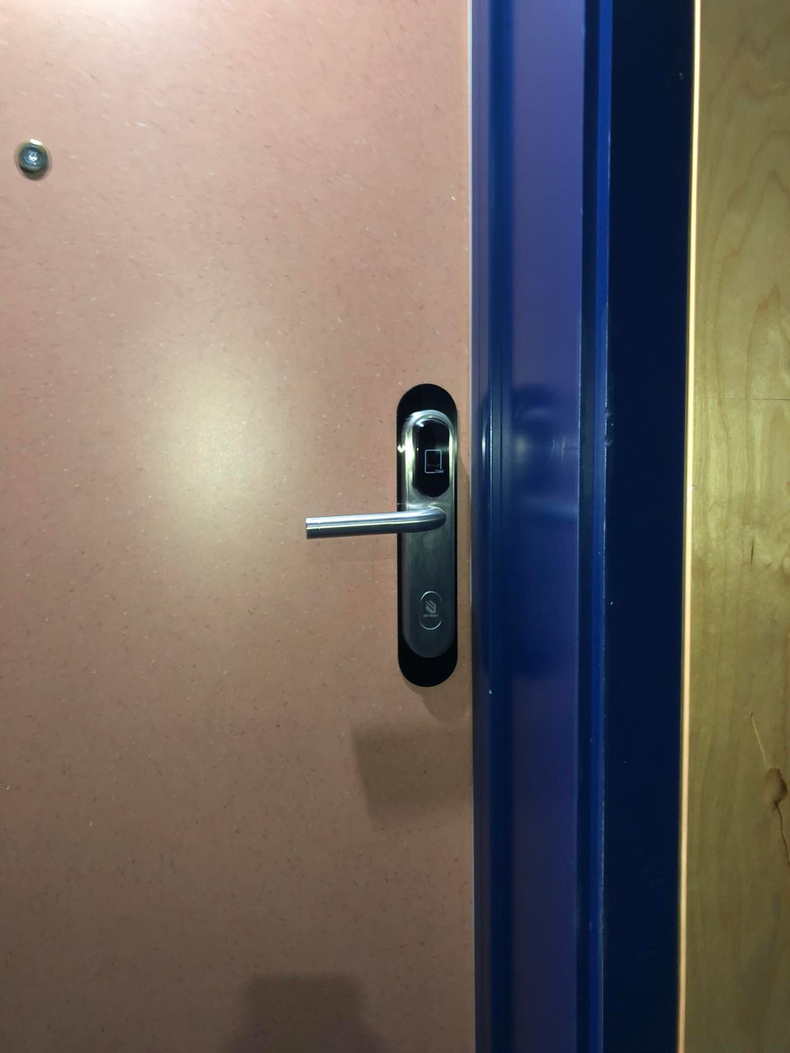 Zewnętrzny zamek elektroniczny z czytnikiem kart na brązowych drzwiach, widoczna niebieska ościeżnica i fragment drewnianej ściany w tle.