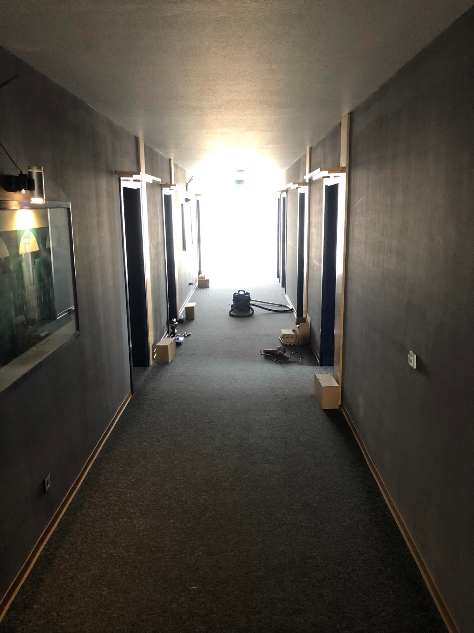 Korytarz w trakcie remontu: szare ściany, ciemna wykładzina, przygotowane ościeżnice, odkurzacz przemysłowy i narzędzia na podłodze.