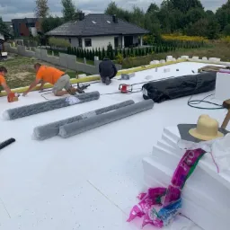 Prace dekarskie: trzech dekarzy w pomarańczowych koszulkach montuje izolację na płaskim dachu domu jednorodzinnego, widoczne rolki papy, styropian, narzędzia i akcesoria dekarskie.