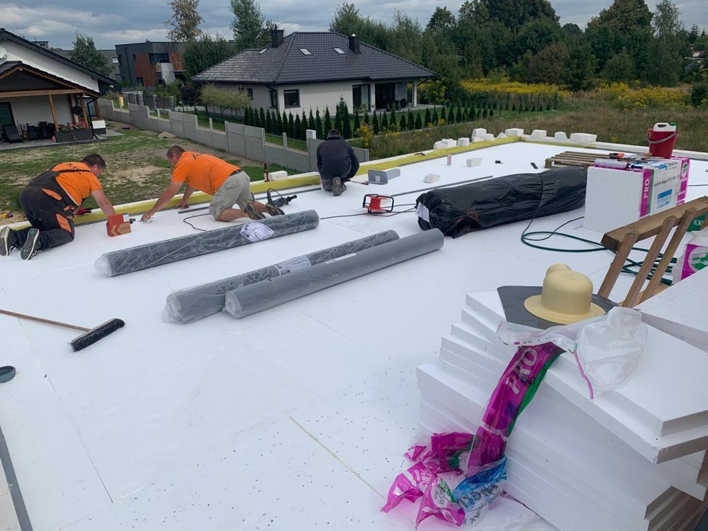 Prace dekarskie: trzech dekarzy w pomarańczowych koszulkach montuje izolację na płaskim dachu domu jednorodzinnego, widoczne rolki papy, styropian, narzędzia i akcesoria dekarskie.