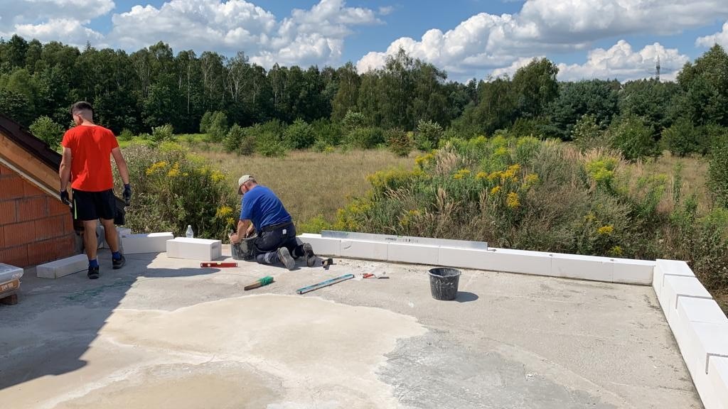 Dwóch pracowników na dachu budynku w trakcie montażu bloczków z betonu komórkowego, z widokiem na zarośla i las w tle, jeden z nich przygotowuje zaprawę w wiadrze.