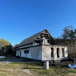 Niedokończony dom z białych bloczków, z częściowo położonym dachem z ciemnej dachówki, otoczony lasem, z widocznymi rusztowaniami i beczką oleju silnikowego Mobil na pierwszym planie.