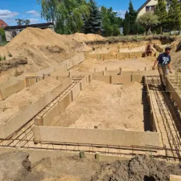 Budowa fundamentów: szalunek z płyty OSB, zbrojenie stalowe, piasek zasypowy, pracownik w koszulce Brooklyn 76 przechodzi po zbrojeniu, w tle drugi pracownik i domy.