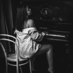 Czarno-białe zdjęcie kobiety siedzącej tyłem na krześle i grającej na starym pianinie z dekoracyjnym portretem w ramie, ubrana w rozpiętą koszulę. Oświetlenie boczne podkreśla fakturę drewna...