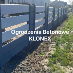 Długi fragment ogrodzenia z prefabrykowanych elementów betonowych, z szarymi słupkami i białymi panelami, widoczny z bliskiej perspektywy wzdłuż drogi gruntowej z trawą.