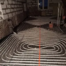 Montaż rur ogrzewania podłogowego w surowym pomieszczeniu z widoczną instalacją elektryczną i oknem w tle.