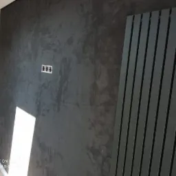 Ściana w ciemnoszarym odcieniu z dekoracyjnym efektem strukturalnym, widoczne gniazdka elektryczne i nowoczesny grzejnik panelowy w kolorze antracytowym.