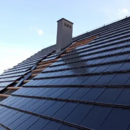 Montaż czarnej dachówki ceramicznej na drewnianej konstrukcji dachu, widoczny komin z szarej blachy i fragment nieba.