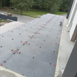 Układanie dużych, szarych płytek na zewnątrz budynku, z użyciem pomarańczowych i czarnych krzyżyków dystansowych, widoczny częściowo wiadro z klejem i otoczenie z drzewami w tle.