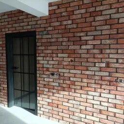 Ściana z czerwonej cegły z czarną, przeszkloną ramą drzwi, widoczne niedokończone instalacje elektryczne i różowe klapki na podłodze.