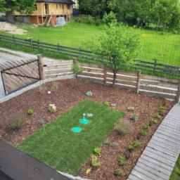 Widok z góry na świeżo założony trawnik z rolki, otoczony korą i roślinami, z widocznymi włazami do systemu nawadniania, ogrodzeniem i domem w tle.