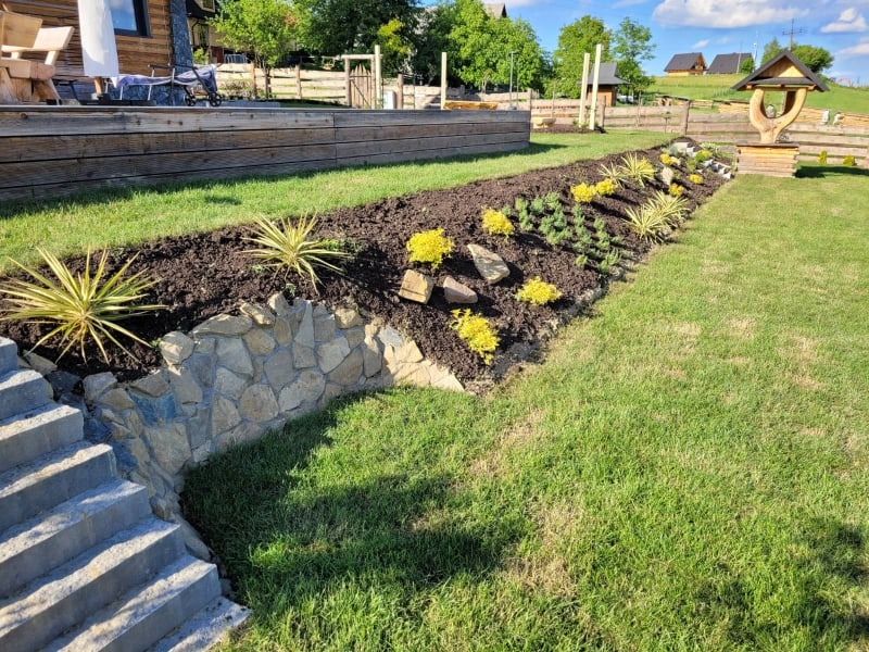 Kaskadowy ogród z kamienną ścianką oporową, świeżo posadzone rośliny ozdobne i trawnik w słoneczny dzień.