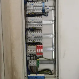 Rozdzielnica elektryczna z zamontowanymi bezpiecznikami, wyłącznikami różnicowoprądowymi i ogranicznikami przepięć, widoczne okablowanie i szyny montażowe w surowym otoczeniu ściany.