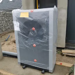 Pompa ciepła powietrze-woda, jeszcze w folii ochronnej, przed budynkiem w Rozłazinie, widoczne naklejki ostrzegawcze i etykieta informacyjna.