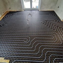 Robert Stencel Usługi Budowlane - Rozłożona instalacja ogrzewania podłogowego na płycie OSB, widoczne białe rurki ułożone w pętle na czarnej macie z żółtymi punktami, w tle okno i surowe ściany.