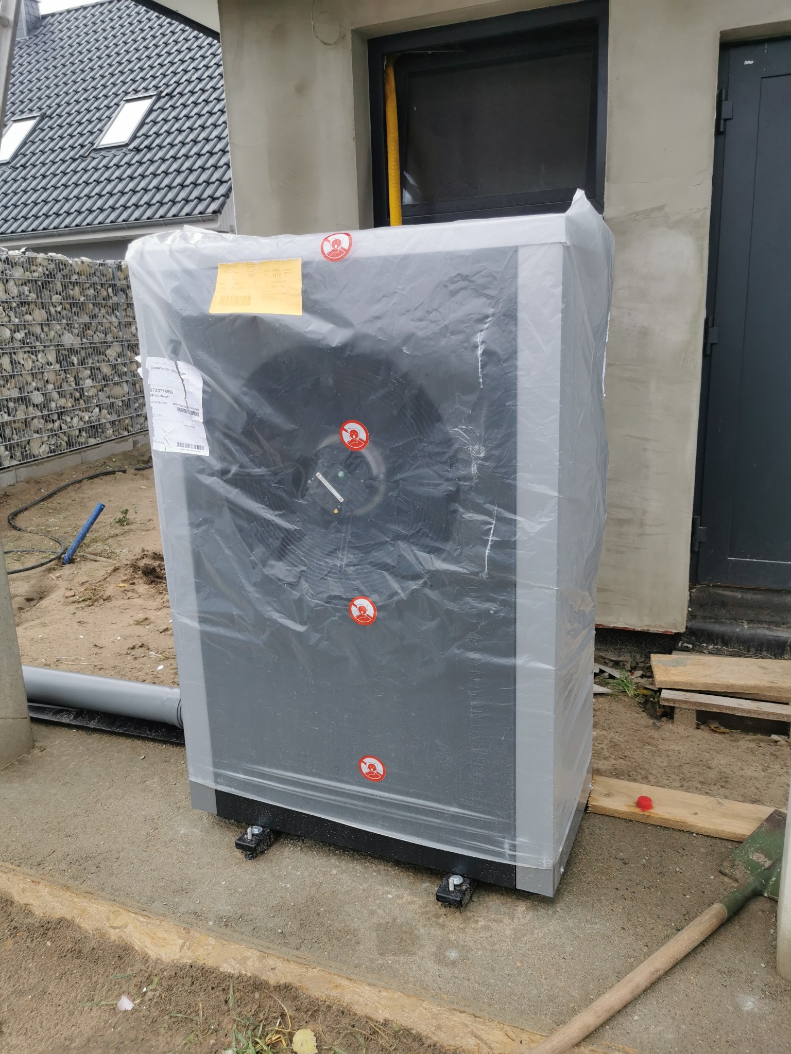 Pompa ciepła powietrze-woda, jeszcze w folii ochronnej, przed budynkiem w Rozłazinie, widoczne naklejki ostrzegawcze i etykieta informacyjna.
