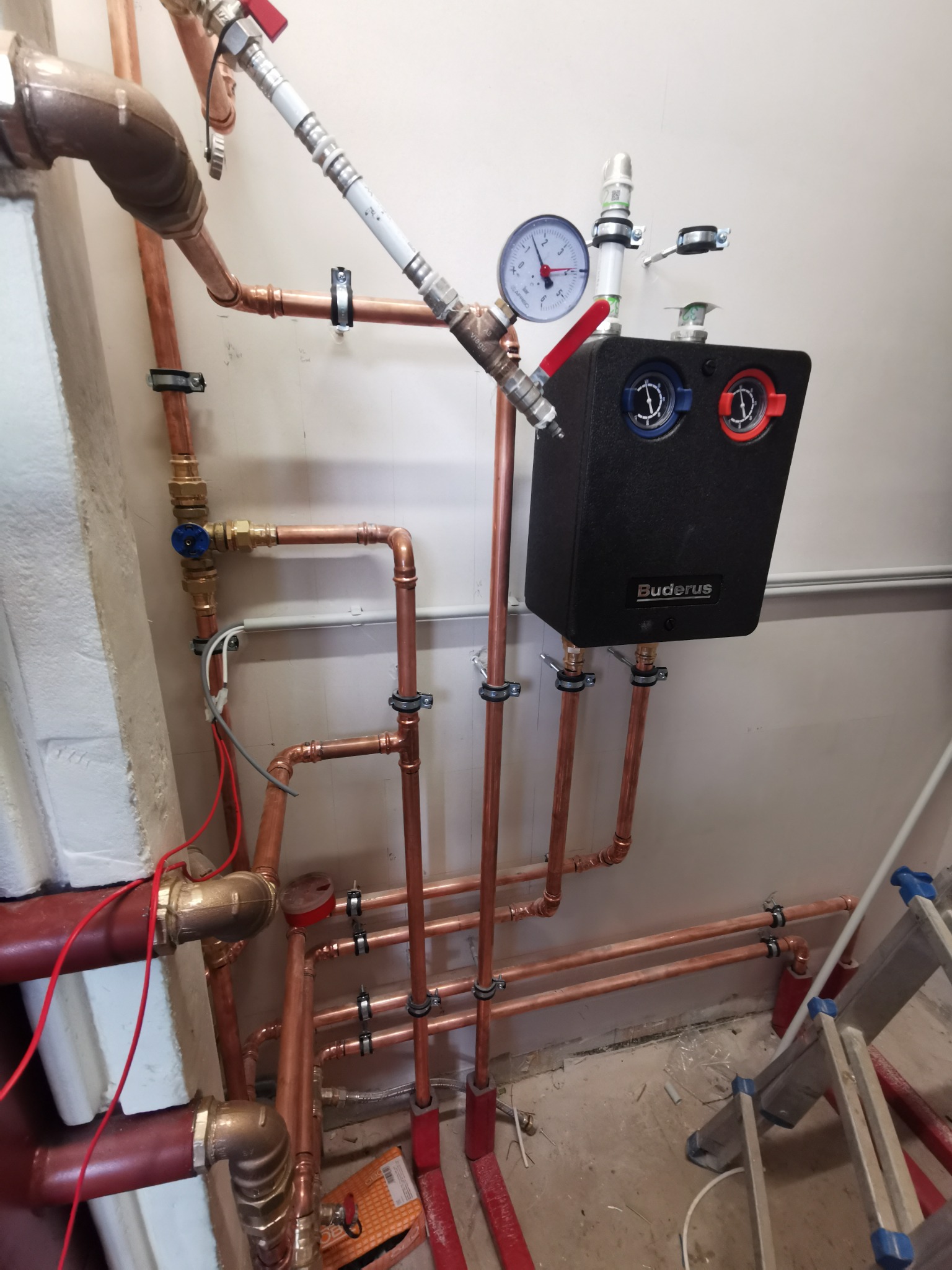 Kompleksowa instalacja grzewcza z miedzianymi rurami, manometrem i czarnym modułem Buderus, widoczna drabina malarska.