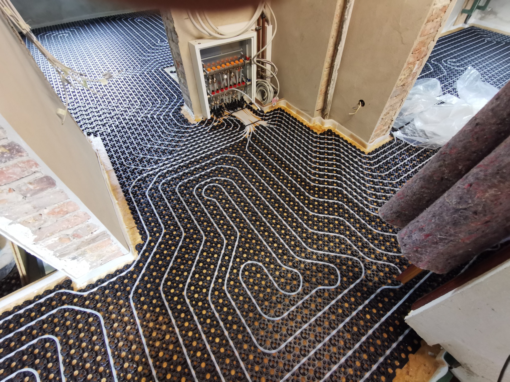 Widok z góry na rozłożone rury ogrzewania podłogowego na czarnej macie z żółtymi punktami, z szafką rozdzielczą w tle i fragmentem ściany w stanie surowym.