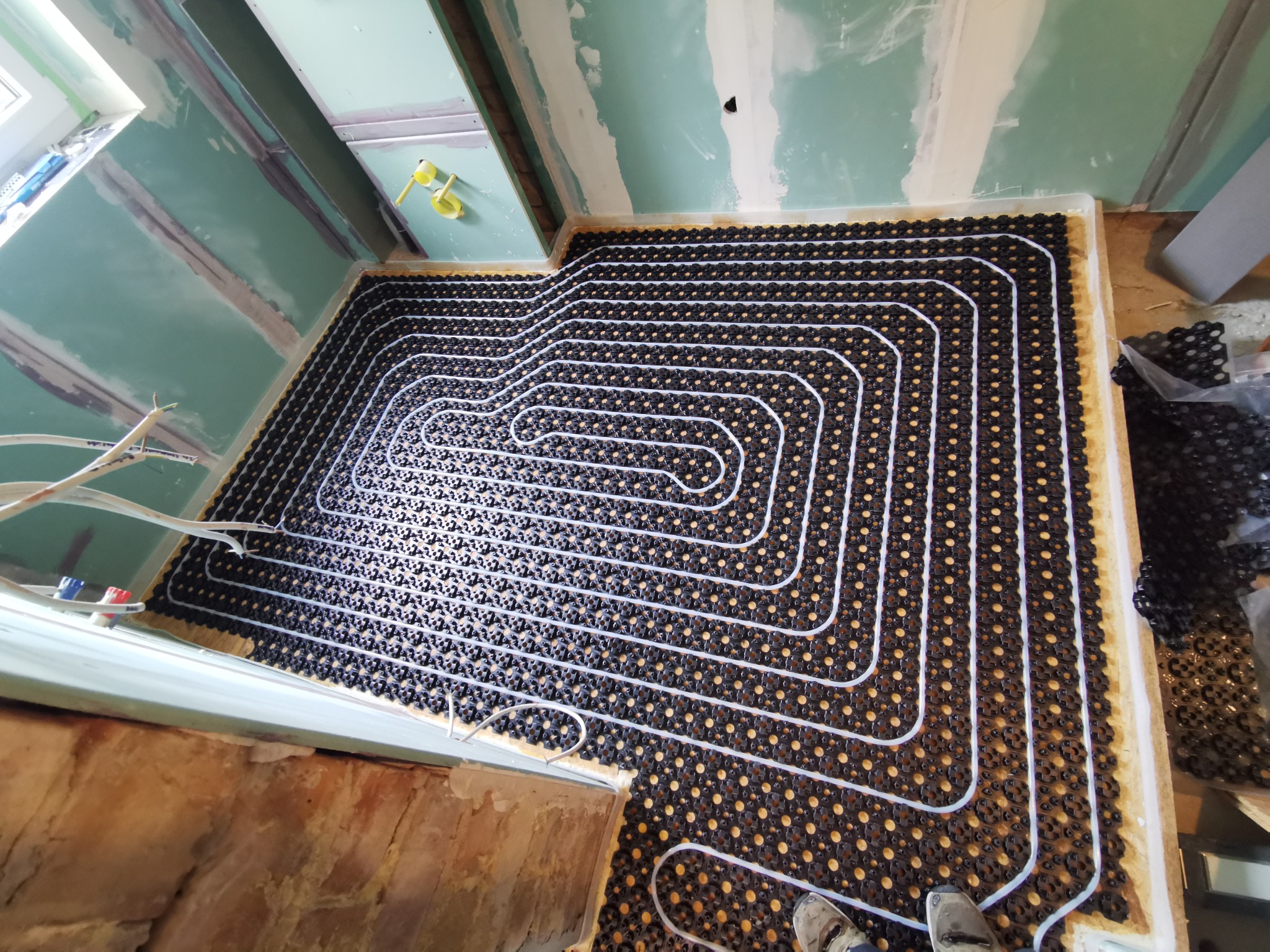 Rozłożona instalacja ogrzewania podłogowego na czarnej macie z wypustkami, widoczne białe rurki, ściany w trakcie wykończenia gipsowego, perspektywa z góry.
