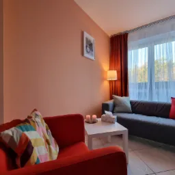 Pokój terapeutyczny z czerwonym fotelem z wzorzystą poduszką, szarą sofą z poduszkami, białym stolikiem z zapalonymi świecami i pudełkiem chusteczek, lampą podłogową i obrazem na ścianie...