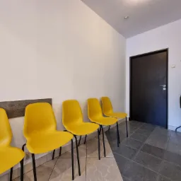 Poczekalnia z rzędem żółtych krzeseł i wieszakiem na ubrania obok ciemnych drzwi.