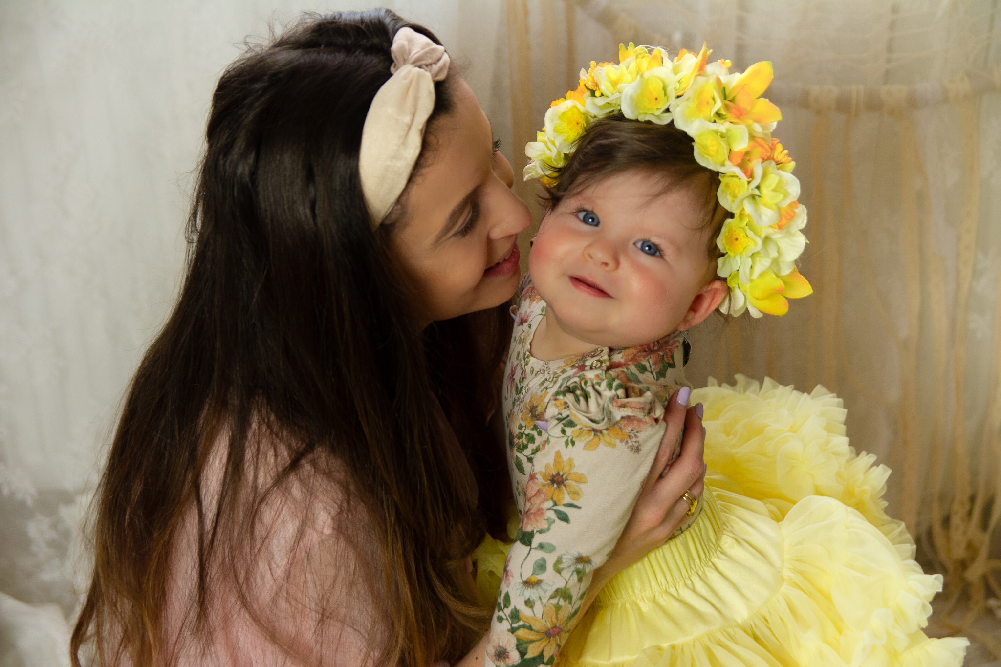 Matka przytula roześmianą córeczkę z niebieskimi oczami i wiankiem z żółtych kwiatów na głowie, ubrana w kwieciste body i żółtą, falbaniastą spódniczkę.