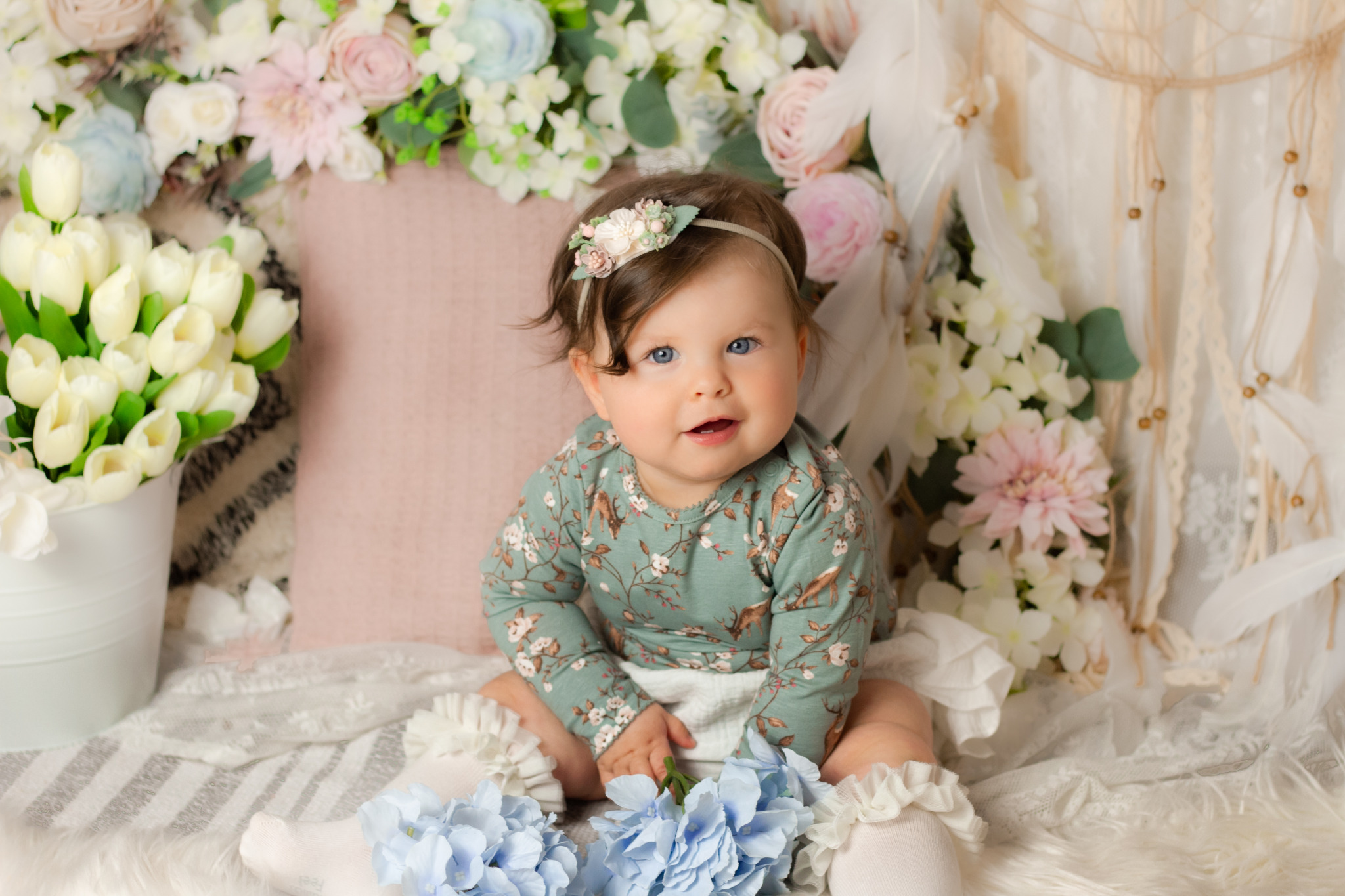 Uśmiechnięta niemowlęca dziewczynka w zielonym body z motywem zwierzęcym i kwiatową opaską na głowie, siedząca na futrzanym kocyku w otoczeniu kwiatów i dekoracji boho, w studio fotograficznym.