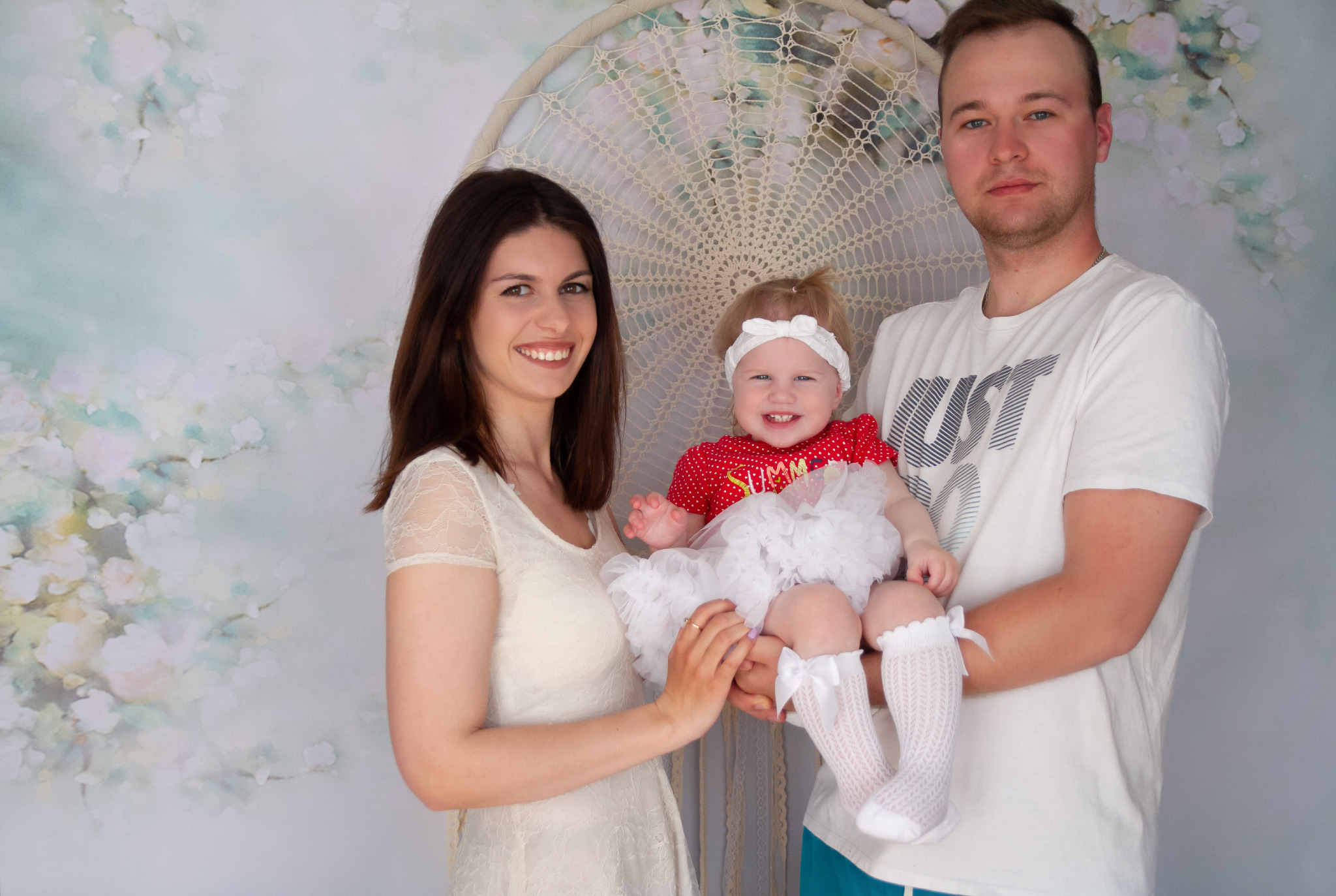 Uśmiechnięta rodzina z małą dziewczynką w czerwonej sukience i białej tutu, pozująca przed dekoracyjnym łapaczem snów i kwiecistym tłem.