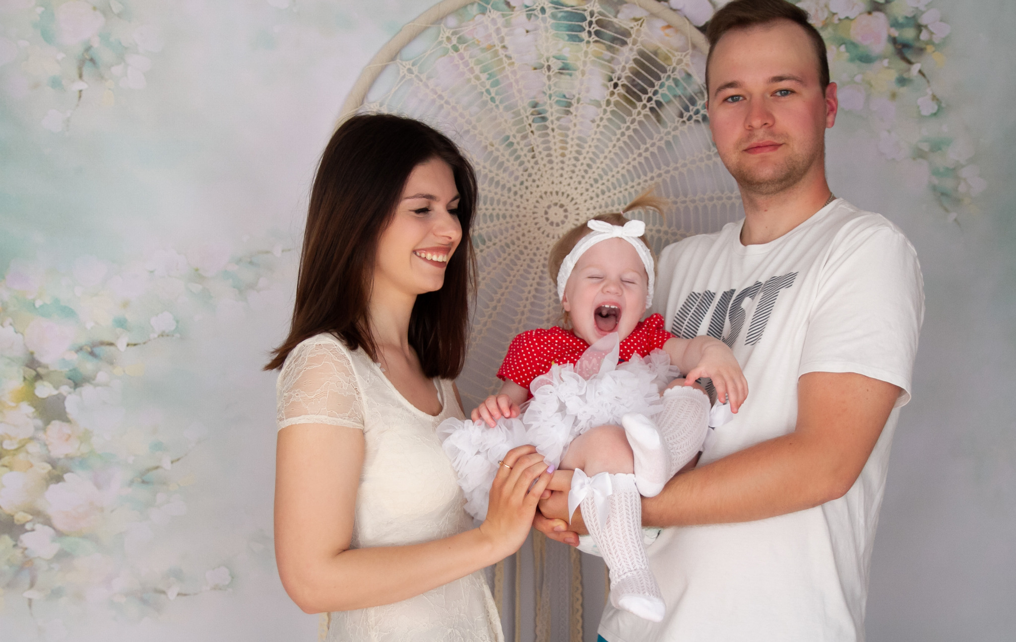Uśmiechnięta mama, tata trzymający na rękach roześmianą dziewczynkę w czerwonej sukience w białe kropki i białej tiulowej spódniczce, na tle dekoracyjnej makramy.
