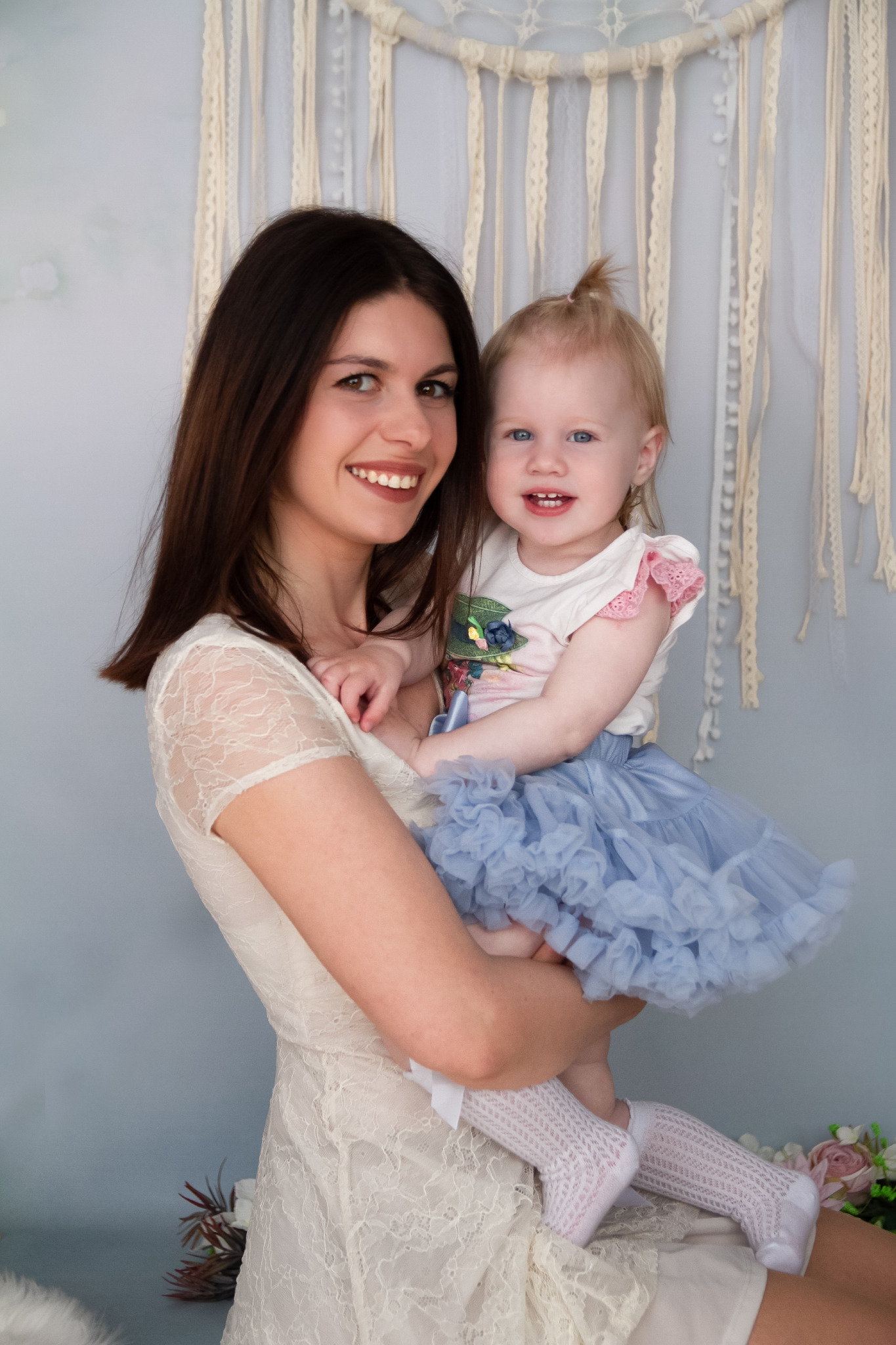 Portret uśmiechniętej kobiety w koronkowej sukience trzymającej na rękach roześmianą dziewczynkę w tiulowej spódniczce i ażurowych podkolanówkach, na tle dekoracji w stylu boho i pastelowych kwiatów.