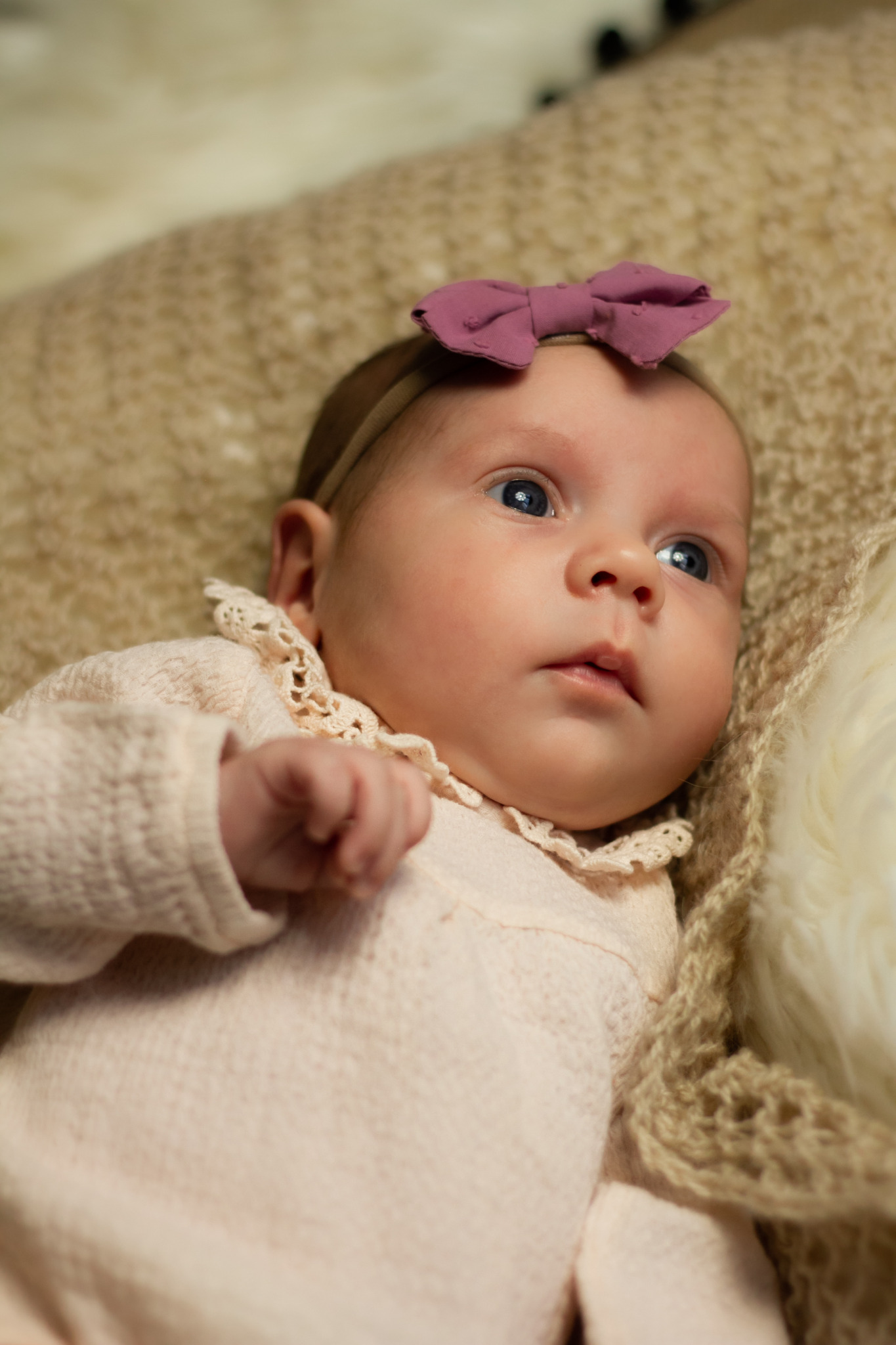 Noworodek leżący na beżowym pledzie, ubrany w różowy sweterek z koronkowym kołnierzykiem i fioletową kokardką na głowie, patrzy w górę.